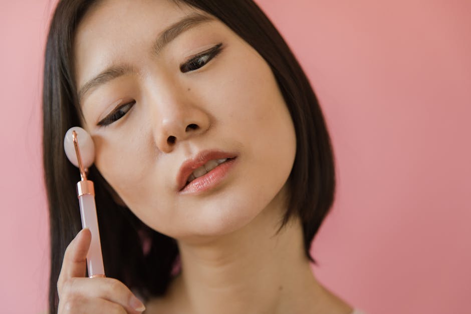 Close-up of an Asian woman using a jade roller for facial massage against a pink background.