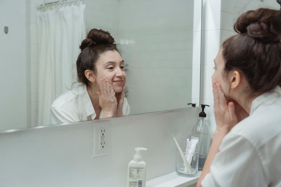 Woman performing skincare routine in a bathroom, applying face cream while looking into a mirror.