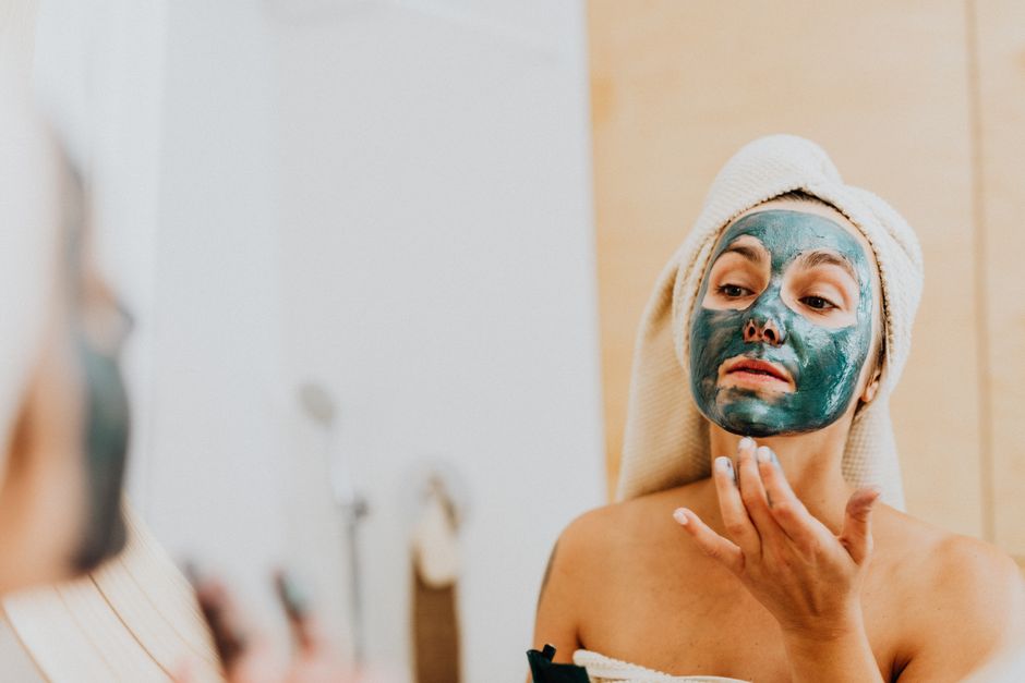 A woman applies a green face mask while looking at her reflection in the bathroom mirror.