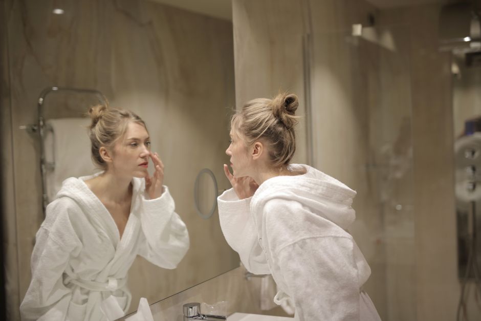 A woman in a bathrobe applies facial cream, reflected in a bathroom mirror, focusing on skincare.