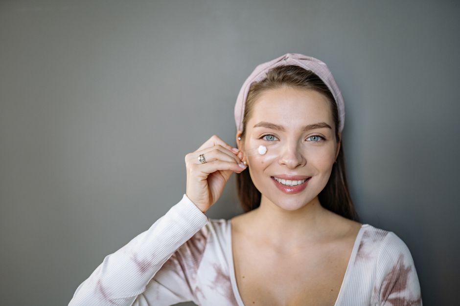 A woman with gray eyes and headband applying skincare cream indoors, smiling.