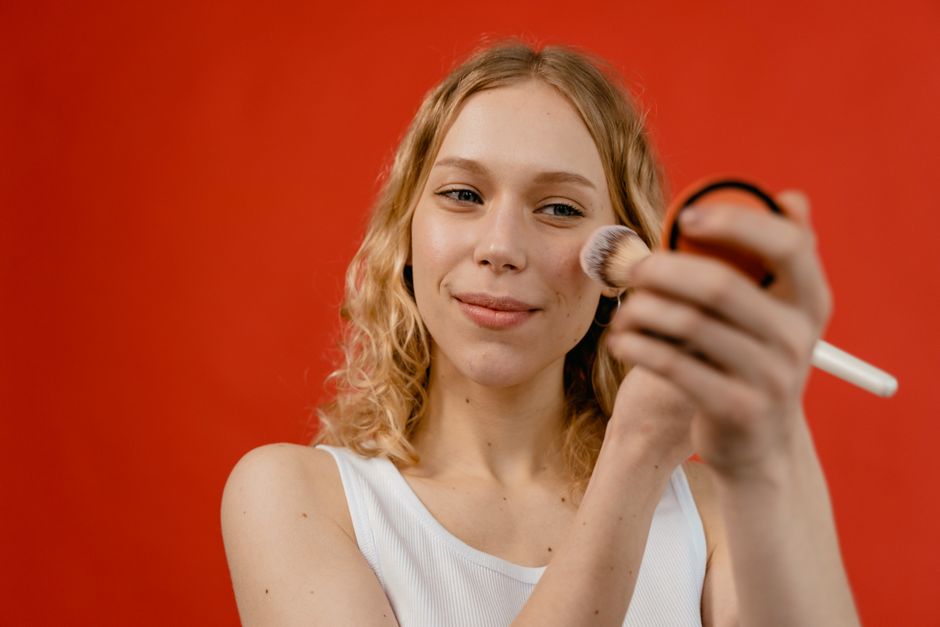 Woman applying makeup with a brush against a vibrant red backdrop, showcasing beauty and skincare concepts.