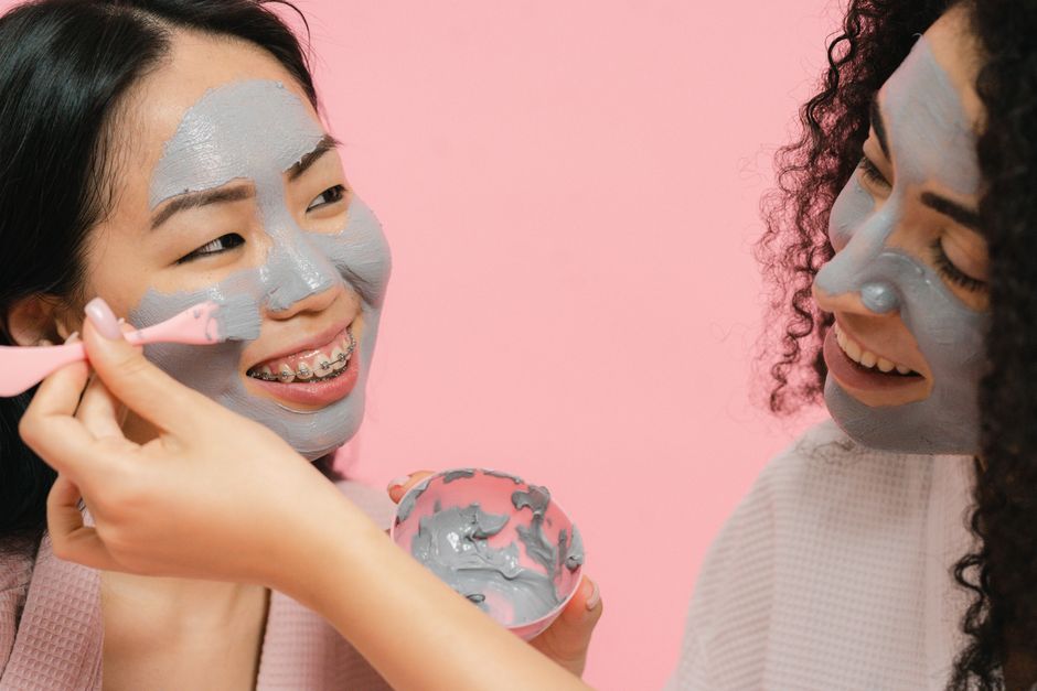 Two women applying gray clay facial masks, smiling and enjoying a relaxing skincare session.