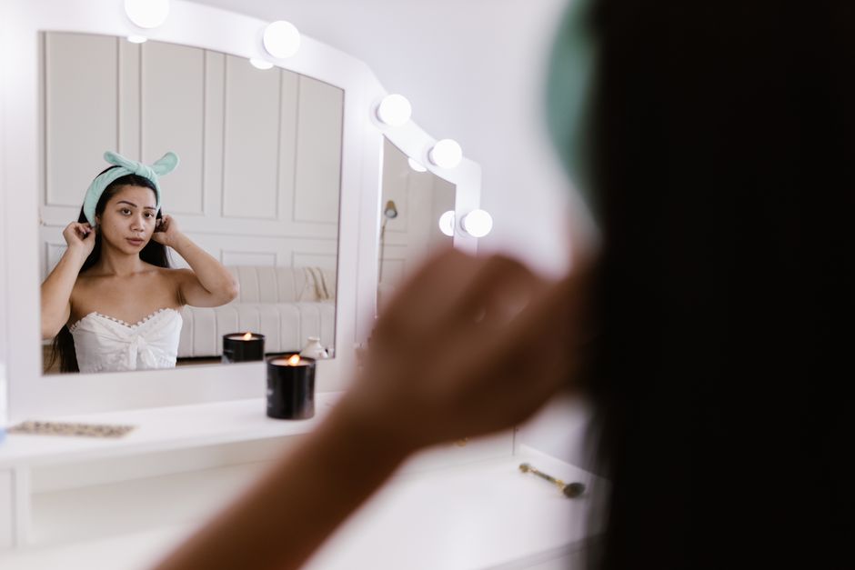 Young woman adjusts hairband at vanity mirror with candles, in serene home setting.