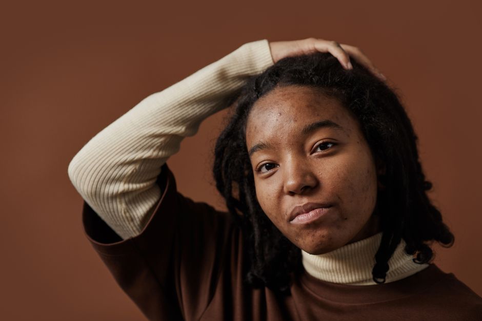 A close-up portrait of a woman with dreadlocks, posing against a brown background.
