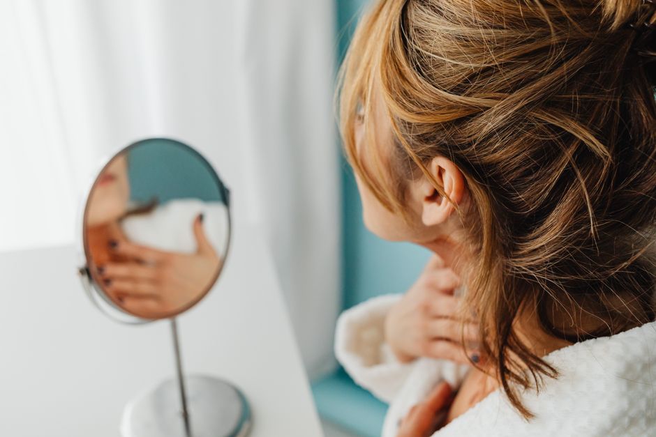 Woman in bathrobe applies skincare while looking in a mirror, highlighting self-care routine.