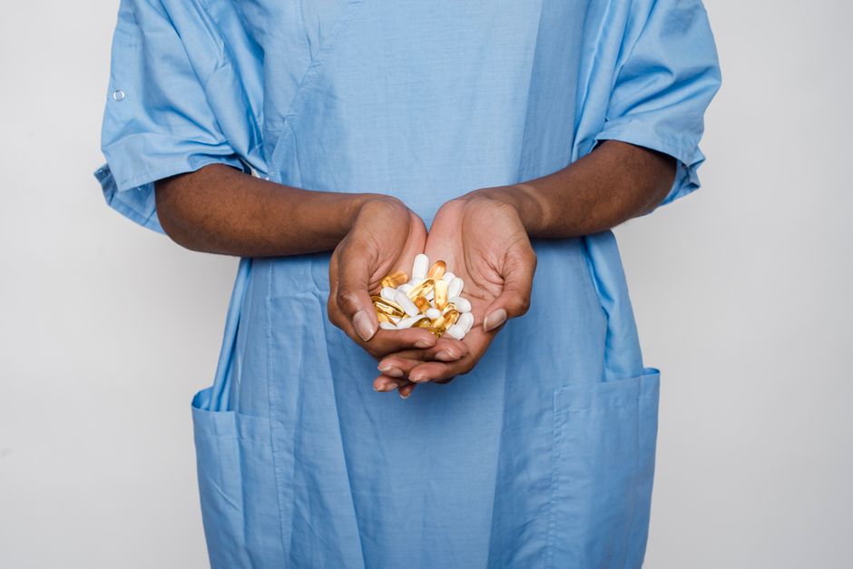 Crop faceless African American patient in blue medical robe demonstrating white and yellow pills in hands on white background in light studio