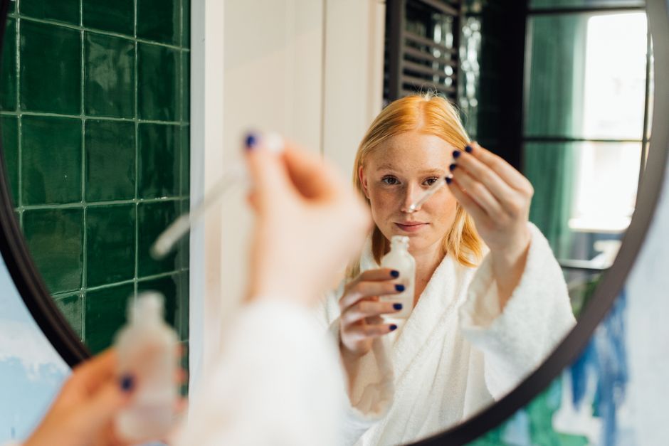 Adult woman applying skincare serum while wearing a bathrobe in front of a bathroom mirror.