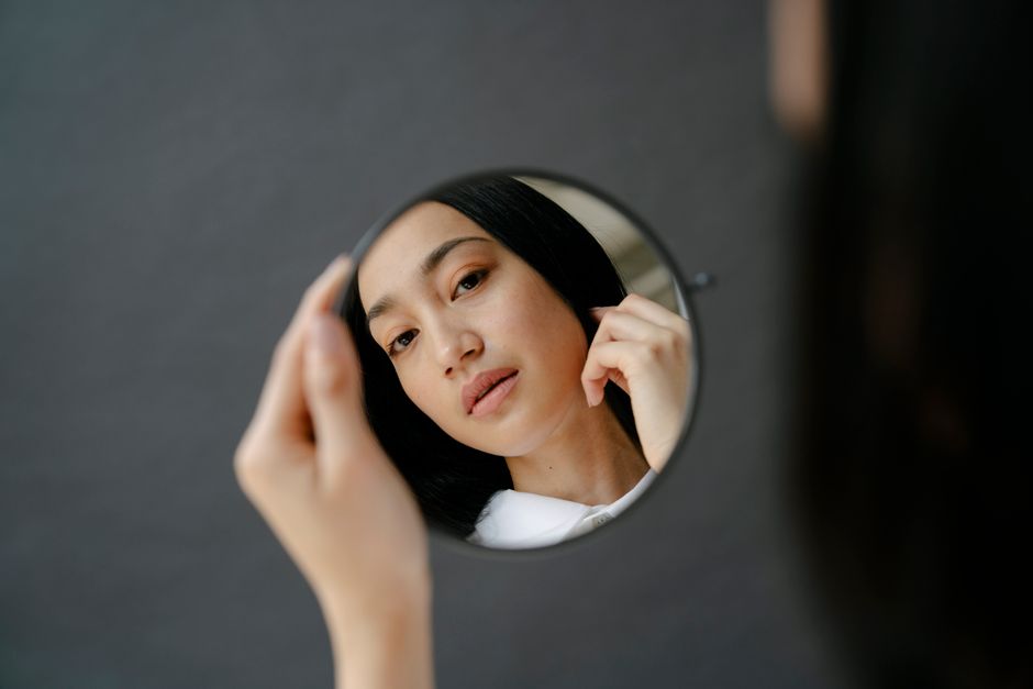 From above of crop attractive Asian female with black hair looking at reflection in round mirror in hand in light room