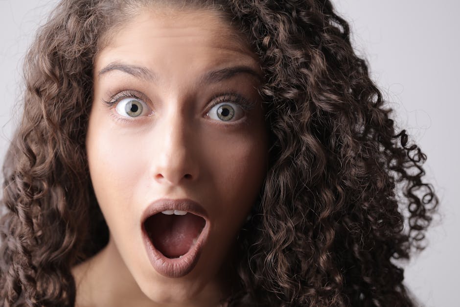 A close-up portrait of a surprised woman with striking eyes and curly hair against a neutral background.