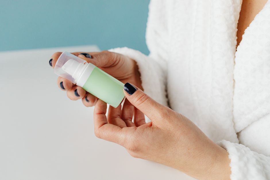 A person in a bathrobe holding a skincare product, focusing on hands and bottle.