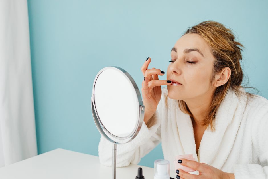 Adult woman in a white bathrobe applying lip care product while looking in a mirror, exemplifying self-care.