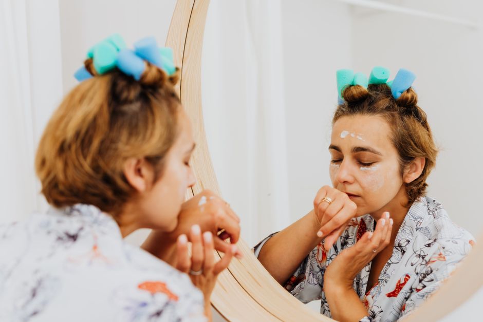 A woman in curlers applies skincare cream while looking in a mirror, reflecting self-care routines.
